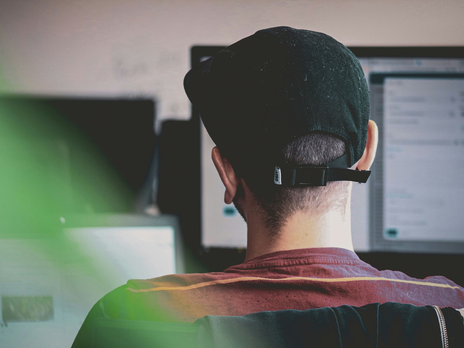 Person mit schwarzer Kappe sitzt vor Computerbildschirmen und arbeitet konzentriert.