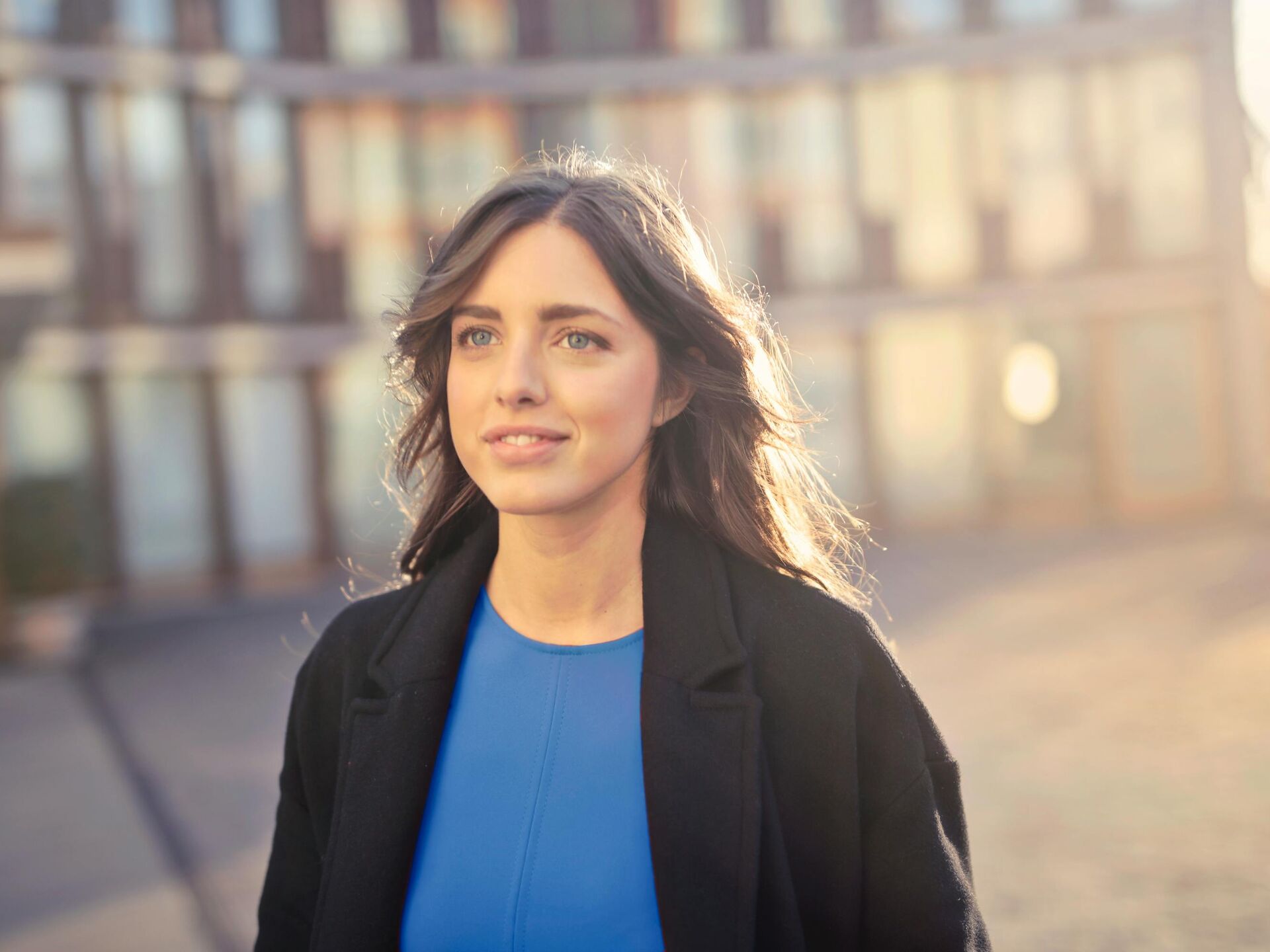 Junge Frau blickt lächelnd nach vorn, im Hintergrund ein modernes Gebäude in warmem Sonnenlicht.