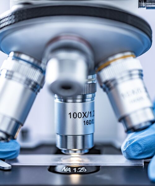 Nahaufnahme eines Mikroskops, unter dem eine Probe mit blauer Handschuhhand positioniert wird.