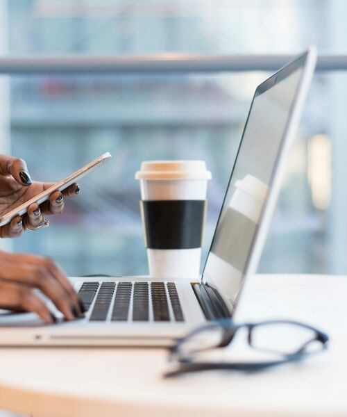 Person arbeitet mit Laptop und Smartphone an einem Tisch, daneben stehen eine Brille und ein Kaffeebecher.