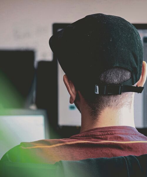 Person mit schwarzer Kappe sitzt vor Computerbildschirmen und arbeitet konzentriert.