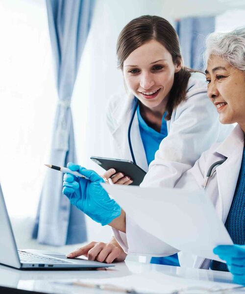 Zwei Ärztinnen betrachten gemeinsam Unterlagen und einen Laptop und besprechen medizinische Daten.