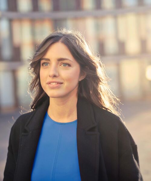 Junge Frau blickt lächelnd nach vorn, im Hintergrund ein modernes Gebäude in warmem Sonnenlicht.